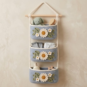 May include: A three-tiered hanging storage organizer made of blue crocheted fabric. Each basket features a white and yellow daisy applique with green leaves. The baskets are suspended by white braided ropes from a horizontal wooden dowel rod.