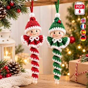 May include: Two hanging ornaments featuring crocheted figures with wooden bead heads and spiral bodies. One figure is red and white, the other is green and white. Both have white collars, matching hats, and are suspended by yarn loops from a wooden branch.