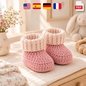 Könnte beinhalten: Ein Paar handgestrickte Babyschuhe mit einer strukturierten, altrosa Basis und einem dicken, gerippten Bündchen in Creme. Die Schuhe sind in einem dichten Häkelmuster gefertigt, was ihnen ein gemütliches Aussehen mit abgerundeter Zehenpartie verleiht.