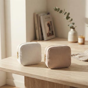 May include: Two square-shaped zippered pouches made of a textured corduroy fabric. One pouch is white and the other is a soft beige color. Both items feature a gold-toned metal zipper pull and a structured, compact design for storage.