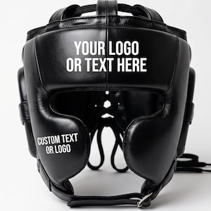 Puede incluir: Un casco de boxeo de cuero negro con diseño acolchado, protección para los oídos y correa para la barbilla. El frente muestra el texto en blanco YOUR LOGO OR TEXT HERE y CUSTOM TEXT OR LOGO, indicando un área personalizable para marca.