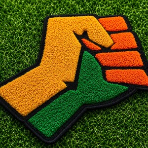 May include: A tufted textile patch featuring a stylised fist design. The artwork is composed of textured yarn in vibrant shades of yellow, orange, and green, with a thick black border outlining the entire shape of the raised fist graphic.