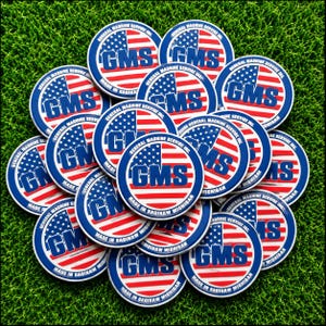 May include: A collection of circular stickers featuring a red, white, and blue American flag design. Each sticker displays the bold blue text GMS in the center, with the words MADE IN SAGINAW MICHIGAN printed along the bottom edge of the border.