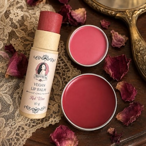 Puede incluir: Un tubo cilíndrico de cartón de bálsamo labial vegano etiquetado Mistress Red Wine 10 g, con una barra de bálsamo rojo intenso, junto a dos latas metálicas redondas que contienen bálsamo labial rojo a juego, rodeado de pétalos de rosa secos y encaje.