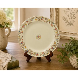 May include: A round ceramic plate with an off-white centre featuring a small floral cluster. The rim is decorated with a continuous pattern of scrolling brown vines, green leaves, and small pink, blue, and yellow flowers on a cream background.