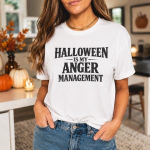 May include: A white short-sleeved t-shirt featuring the black text "HALLOWEEN IS MY ANGER MANAGEMENT" printed in a bold, stylized font across the chest. The garment is paired with blue denim jeans, showcasing a casual and standard fit design.