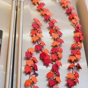 Peut inclure: Trois guirlandes de vigne décoratives verticales composées de feuilles en tissu feutré dans des tons vifs de rouge, d'orange et de cramoisi profond. Chaque vigne possède une tige brune texturée avec de petites feuilles superposées, créant un affichage dense.