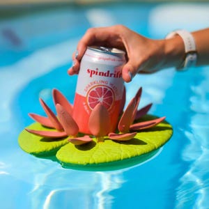 Op de afbeelding: Een blikje Spindrift grapefruit bruiswater geplaatst in een drijvende waterlelie-drankhouder. De houder heeft een felgroene ronde basis met textuuraders en verschillende rechtopstaande roze bloemblaadjes rondom het blikje.