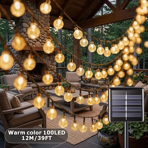 May include: A string of 100 warm-colored LED globe lights measuring 39 feet in length, featuring a black solar panel on a stake. The clear, textured spherical bulbs are arranged on a dark wire, emitting a soft glow for outdoor illumination.