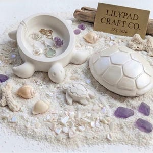 May include: A white turtle-shaped trinket dish with a matching removable shell lid, accompanied by a small turtle figurine. The dish contains two silver necklaces with pendants. A rectangular sign reads LILYPAD CRAFT CO. EST. 2026.