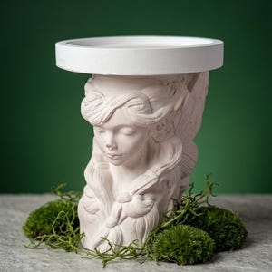 May include: A white matte pedestal stand featuring a sculpted bust of a person with closed eyes and braided hair. The top of the stand is a flat, circular platform. The sculpture is crafted from a solid material with a smooth, light-colored finish.