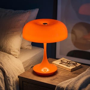 May include: A bright orange table lamp with a smooth, mushroom-shaped shade and a slender, curved stem base. The lamp features a glossy finish and a small metallic accent at the center top of the shade, emitting a warm, soft glow from within.