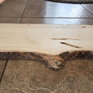 Pu&ograve; includere: Una lastra di legno naturale con un bordo vivo e un colore marrone chiaro. La lastra ha alcuni nodi e una texture ruvida.