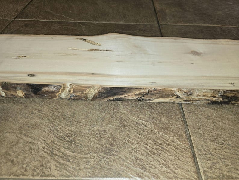 Pu&ograve; includere: Primo piano di una tavola di legno di colore chiaro con un bordo naturale. Il legno ha un colore marrone chiaro e una superficie liscia. Il bordo della tavola &egrave; ruvido e di colore marrone scuro.