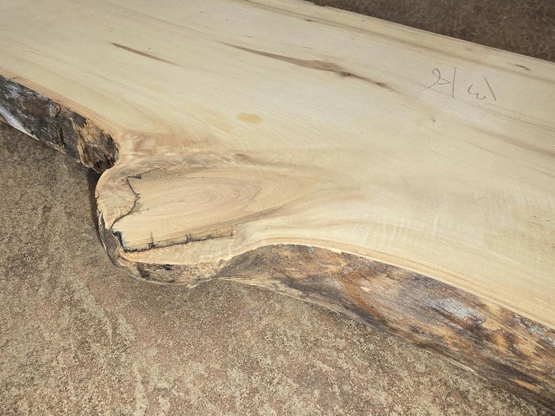 Pu&ograve; includere: Una grande lastra di legno marrone chiaro con un bordo naturale. Il legno ha un colore marrone chiaro con striature marrone scuro. La lastra ha una texture ruvida e naturale. La lastra &egrave; contrassegnata dal testo scritto a mano "2/a1".