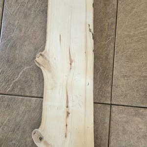 Pu&ograve; includere: Una tavola di legno chiaro con un bordo naturale e nodi. La tavola ha misure scritte a mano, tra cui "15 1/2" e "2 1/8".