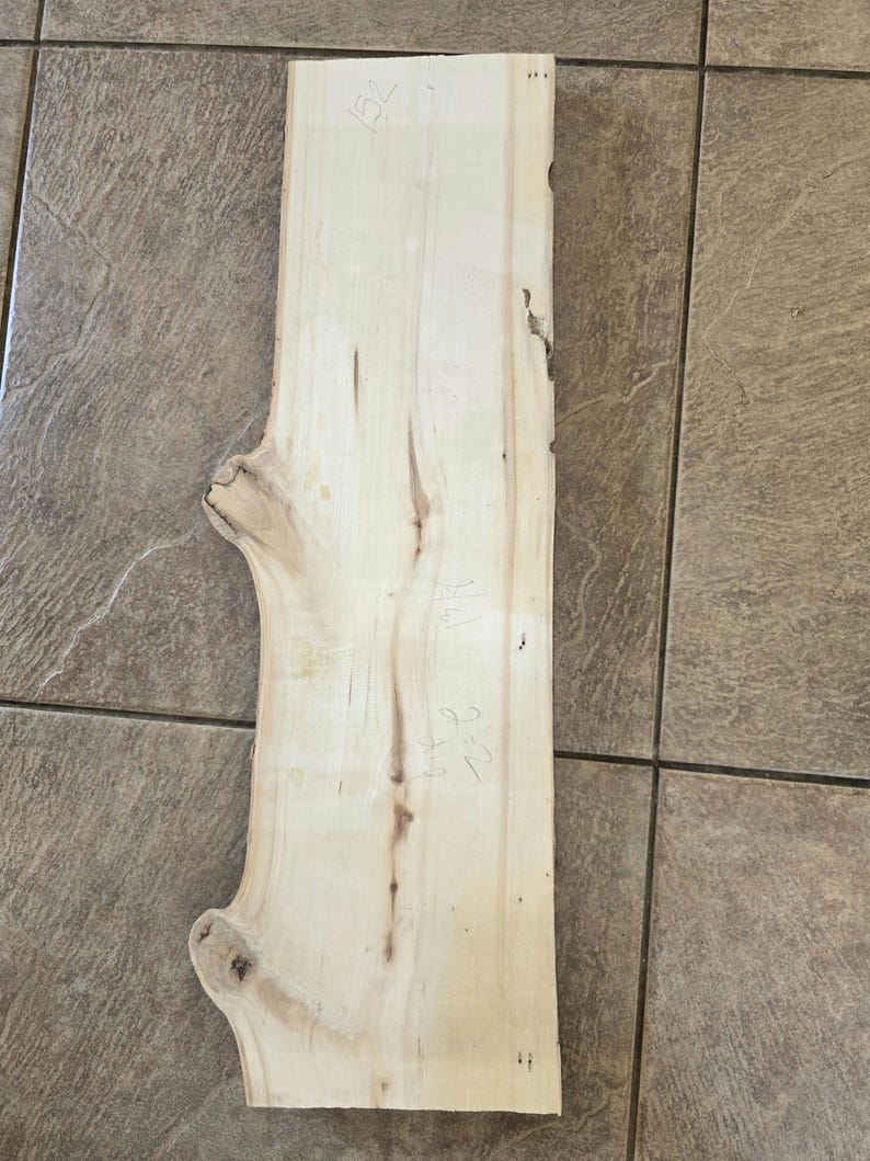 Pu&ograve; includere: Una tavola di legno chiaro con un bordo naturale e nodi. La tavola ha misure scritte a mano, tra cui "15 1/2" e "2 1/8".