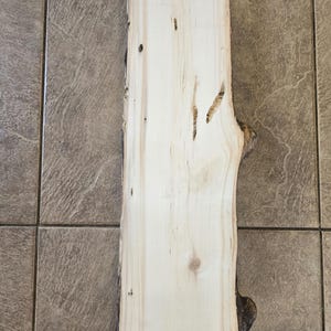 Pu&ograve; includere: Una tavola di legno marrone chiaro con un bordo naturale e alcuni nodi. La tavola &egrave; liscia e ha una venatura del legno chiara.