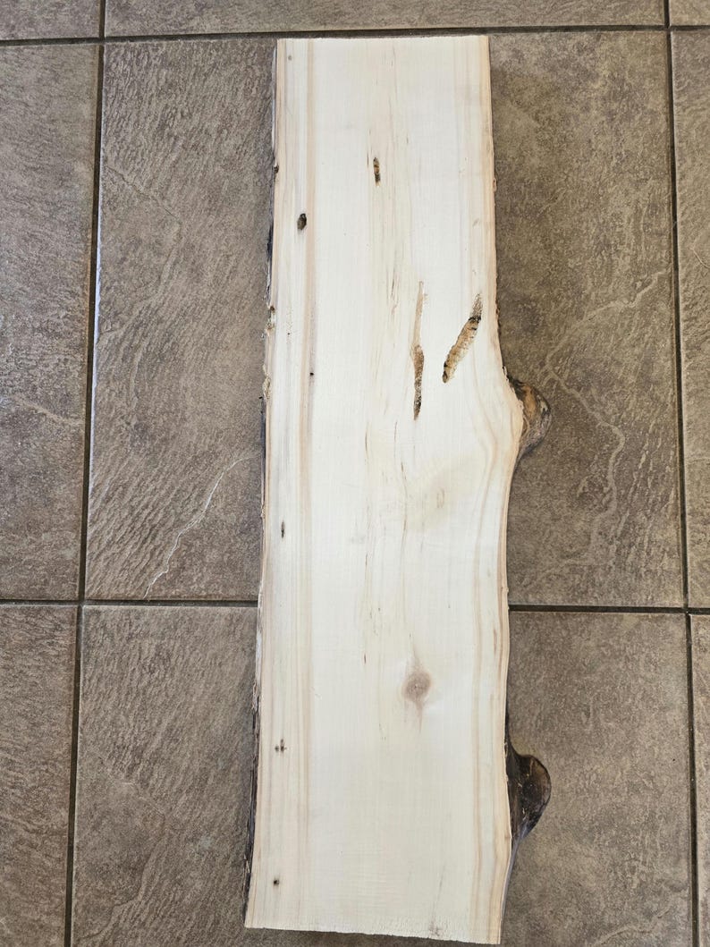 Pu&ograve; includere: Una tavola di legno marrone chiaro con un bordo naturale e alcuni nodi. La tavola &egrave; liscia e ha una venatura del legno chiara.