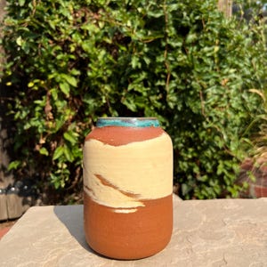 May include: A cylindrical ceramic vase featuring a matte terracotta base, a wide band of cream-colored glaze across the center, and a thin, vibrant turquoise rim at the top opening. The vessel has a smooth, rounded shape with a flat base.