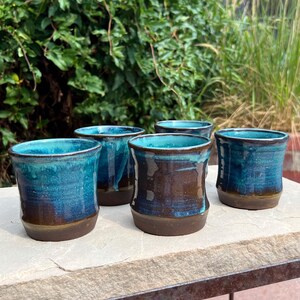 May include: A set of four ceramic cups featuring a two-tone glaze with a deep brown base that transitions into a vibrant turquoise blue at the top. Each cup has a cylindrical shape with a slightly wider rim and a smooth, glossy finish on the exterior.