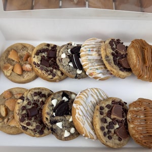 May include: A white cardboard box containing twelve assorted round cookies arranged in two rows. The selection includes cookies topped with chocolate chunks, white chocolate chips, sandwich cookie pieces, white icing drizzles, and caramel drizzles.