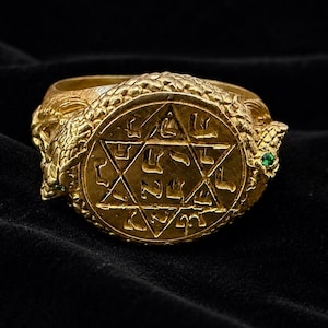 Peut inclure: Une bague en or présentant un médaillon circulaire central avec une étoile à six branches gravée et des caractères hébreux. L'anneau est conçu comme deux serpents entrelacés aux écailles texturées, chacun doté d'une petite pierre verte.