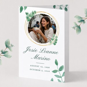 Può includere: Un biglietto commemorativo piegato con una cornice circolare per foto con bordo dorato, decorato con foglie di eucalipto ad acquerello. Il testo recita Josie Leanne Marino, 15 agosto 1998 e 5 ottobre 2026, in caratteri serif verde scuro e nero.