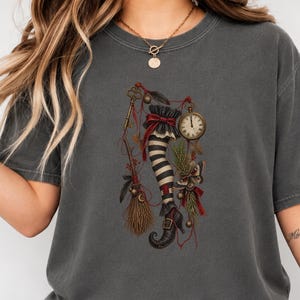 May include: A charcoal grey short-sleeve t-shirt featuring a central graphic of a striped stockinged leg wearing a buckled shoe, surrounded by a vintage pocket watch, a broom, a golden key, pine needles, and a moth, all tied with red ribbons.