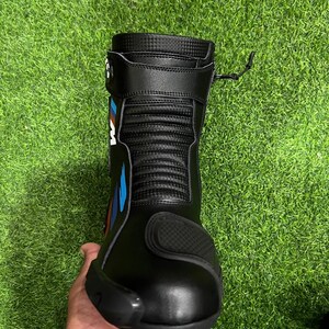 Puede incluir: Una bota de motocicleta negra con parte superior texturizada, puntera reforzada y una correa lateral con un logotipo en azul, rojo y blanco. La bota incluye un cierre de cremallera vertical en el lateral y un panel acanalado flexible en el tobillo.