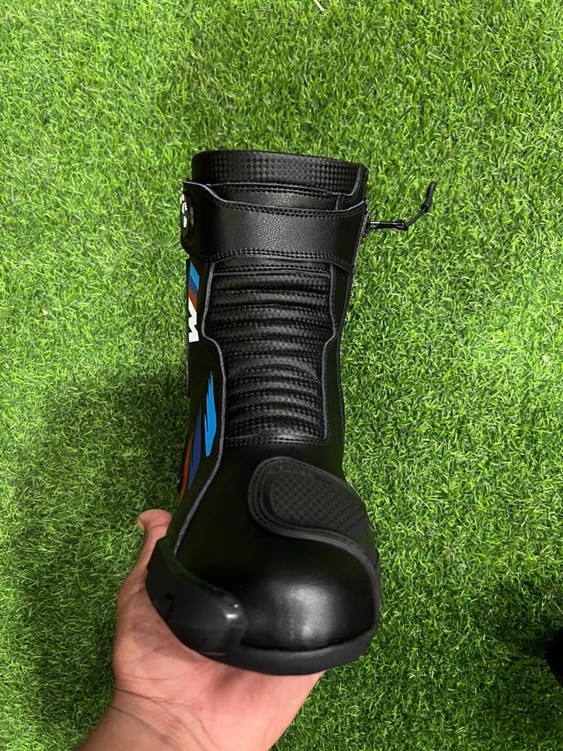 Puede incluir: Una bota de motocicleta negra con parte superior texturizada, puntera reforzada y una correa lateral con un logotipo en azul, rojo y blanco. La bota incluye un cierre de cremallera vertical en el lateral y un panel acanalado flexible en el tobillo.