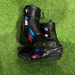 Puede incluir: Un par de botas de moto negras con el logotipo de BMW y la marca M con detalles de pinceladas azules, moradas y rojas. Las botas tienen un dise&ntilde;o de ca&ntilde;a alta con deslizadores de puntera reforzados, paneles texturizados y correas ajustables.