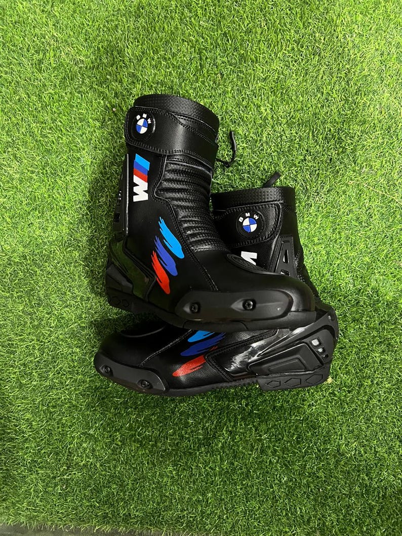 Puede incluir: Un par de botas de moto negras con el logotipo de BMW y la marca M con detalles de pinceladas azules, moradas y rojas. Las botas tienen un dise&ntilde;o de ca&ntilde;a alta con deslizadores de puntera reforzados, paneles texturizados y correas ajustables.