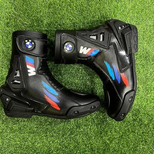 Puede incluir: Un par de botas de moto negras con el logotipo de BMW en la correa del tobillo, la letra M en blanco y detalles de pinceladas azules y rojas. Las botas tienen protectores de tal&oacute;n y puntera de pl&aacute;stico negro reforzado con textura.
