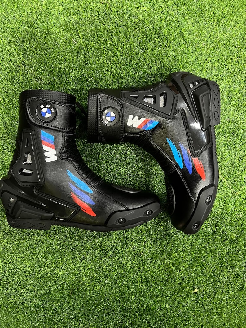 Puede incluir: Un par de botas de moto negras con el logotipo de BMW en la correa del tobillo, la letra M en blanco y detalles de pinceladas azules y rojas. Las botas tienen protectores de tal&oacute;n y puntera de pl&aacute;stico negro reforzado con textura.