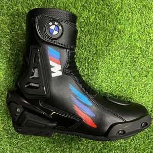 Puede incluir: Una bota de moto negra con el logotipo de BMW en la correa superior, una letra M blanca y detalles de pinceladas en azul, morado y rojo. La bota incluye un protector de tal&oacute;n negro, una puntera reforzada y un cuello con textura.