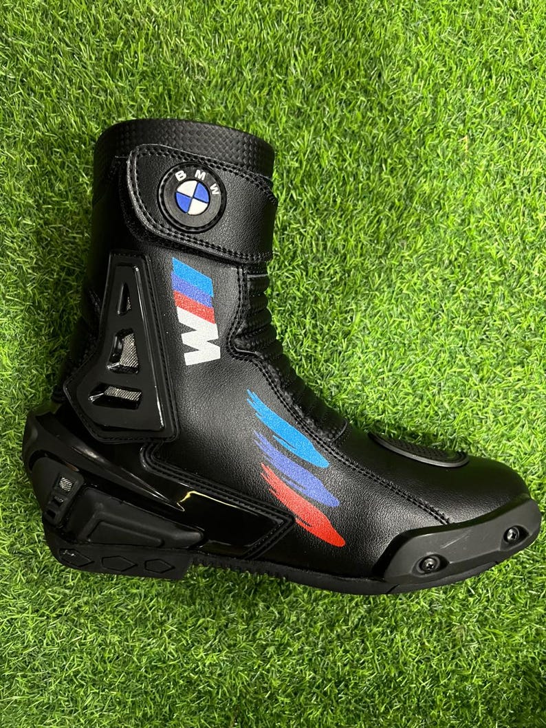 Puede incluir: Una bota de moto negra con el logotipo de BMW en la correa superior, una letra M blanca y detalles de pinceladas en azul, morado y rojo. La bota incluye un protector de tal&oacute;n negro, una puntera reforzada y un cuello con textura.