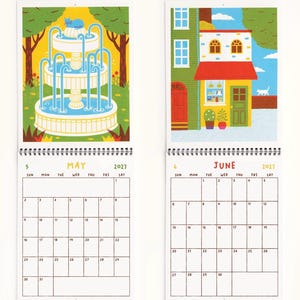May include: A 2027 wall calendar featuring two open pages for May and June. May displays a blue cat on a tiered fountain in a park. June shows a colorful storefront with a red awning and a white cat walking on a brick wall. Both pages include a grid calendar.