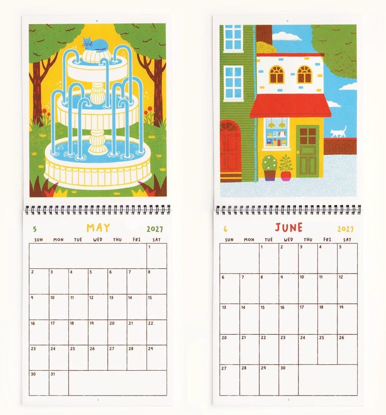 May include: A 2027 wall calendar featuring two open pages for May and June. May displays a blue cat on a tiered fountain in a park. June shows a colorful storefront with a red awning and a white cat walking on a brick wall. Both pages include a grid calendar.