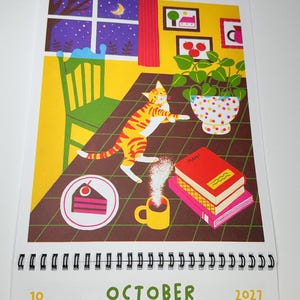 May include: A wall calendar for October 2027 featuring an illustration of an orange tabby cat on a table with a slice of cake, a steaming yellow mug, a stack of books, and a potted plant. The background shows a window at night.