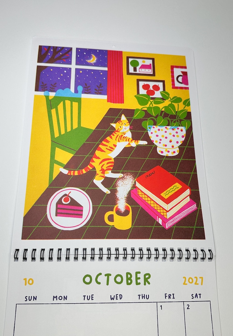 May include: A wall calendar for October 2027 featuring an illustration of an orange tabby cat on a table with a slice of cake, a steaming yellow mug, a stack of books, and a potted plant. The background shows a window at night.