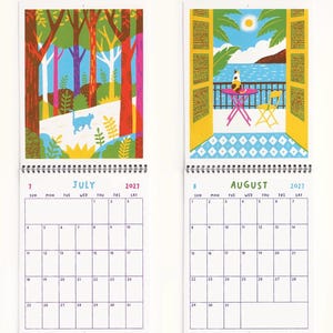 May include: A 2027 wall calendar featuring two open pages for July and August. July displays a colorful forest illustration with a blue cat. August shows a balcony view with a cat, a pink table, and a yellow chair overlooking the sea.