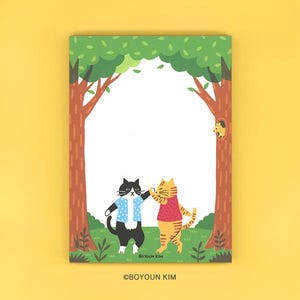 Può includere: Un blocco note rettangolare con un'illustrazione di una foresta e due gatti che si battono il cinque al centro. Un gatto è bianco e nero con una camicia blu a pois, l'altro è un gatto tigrato arancione con una maglietta rossa. Testo: BOYOUN KIM.