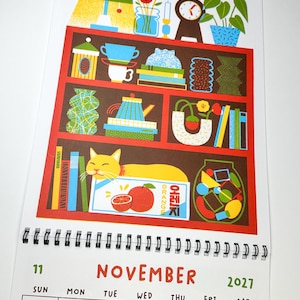 May include: A November 2021 wall calendar page featuring a colorful illustration of a red bookshelf filled with various objects including books, vases, a clock, a lamp, and a yellow cat resting on a box labeled ORANGE in English and Korean.