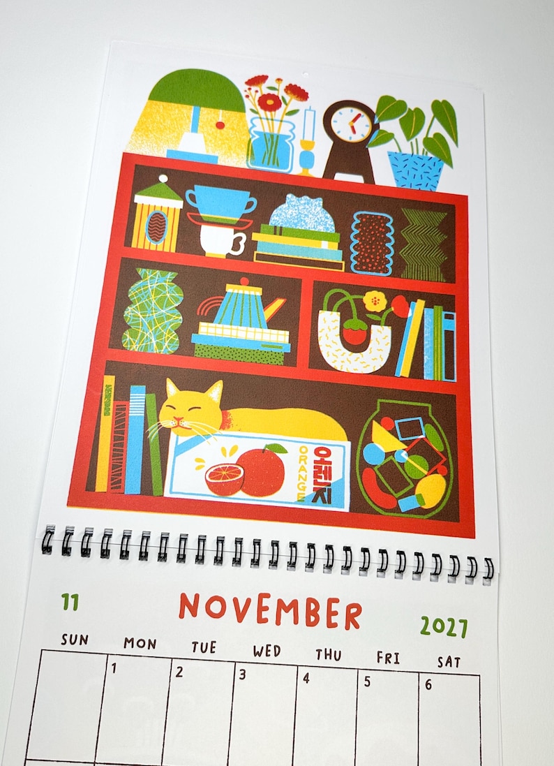 May include: A November 2021 wall calendar page featuring a colorful illustration of a red bookshelf filled with various objects including books, vases, a clock, a lamp, and a yellow cat resting on a box labeled ORANGE in English and Korean.