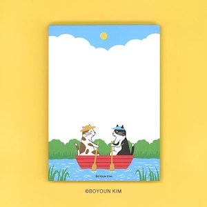 May include: A rectangular notepad featuring a graphic illustration of two cats in a red rowboat on blue water. One cat wears a yellow hat, the other a blue cap. The scene includes green bushes, a blue sky with white clouds, a yellow sun, and the text BOYOUN KIM.