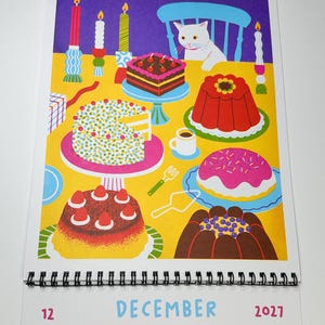 May include: A December 2027 wall calendar page featuring a colorful illustration of a white cat sitting at a yellow table filled with various cakes, desserts, and lit candles. The calendar grid below displays the month name and dates.