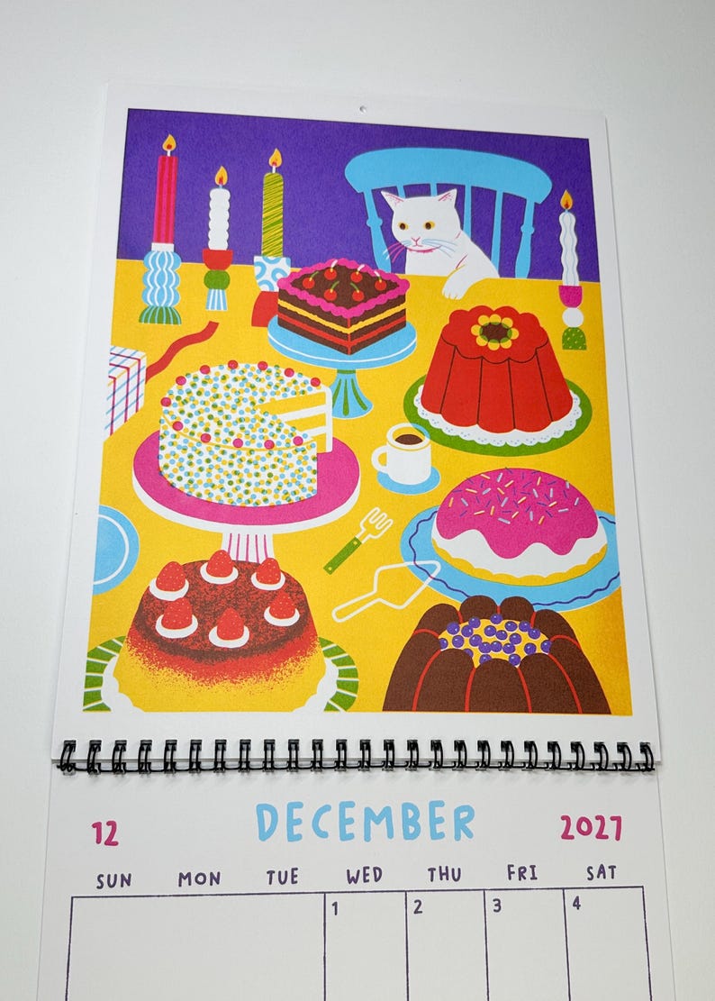 May include: A December 2027 wall calendar page featuring a colorful illustration of a white cat sitting at a yellow table filled with various cakes, desserts, and lit candles. The calendar grid below displays the month name and dates.