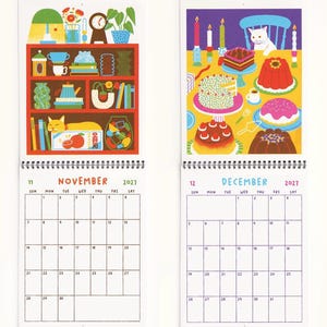 May include: A 2027 wall calendar featuring two open pages for November and December. November displays a red shelf with a cat and various objects. December shows a white cat at a table with cakes and candles. Both pages include a grid calendar.