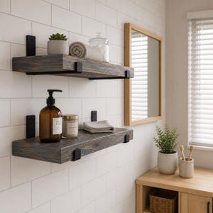 May include: Two dark stained wooden floating shelves mounted on a white tiled wall with black metal brackets. The top shelf holds a small potted succulent, a rolled beige towel, and a glass jar. The bottom shelf holds a brown soap dispenser.